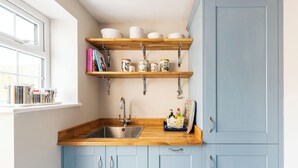 Een keuken met houten aanrecht, een spoelbak en blauwe kastjes. Boven de spoelbak staan planken met potten en boeken.