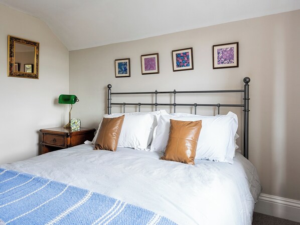 A neatly made bed with white linens and brown pillows, a green desk lamp, and a framed mirror on the wall. - Charming cottage near lovely Stamford with cosy open fire and a rural location. (Stamford, near Easton-on-the-Hill)