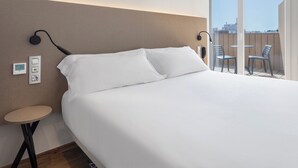 Habitació d'hotel amb un llit gran, una taula rodona petita, una làmpada penjada a la paret i un balcó amb cadires.