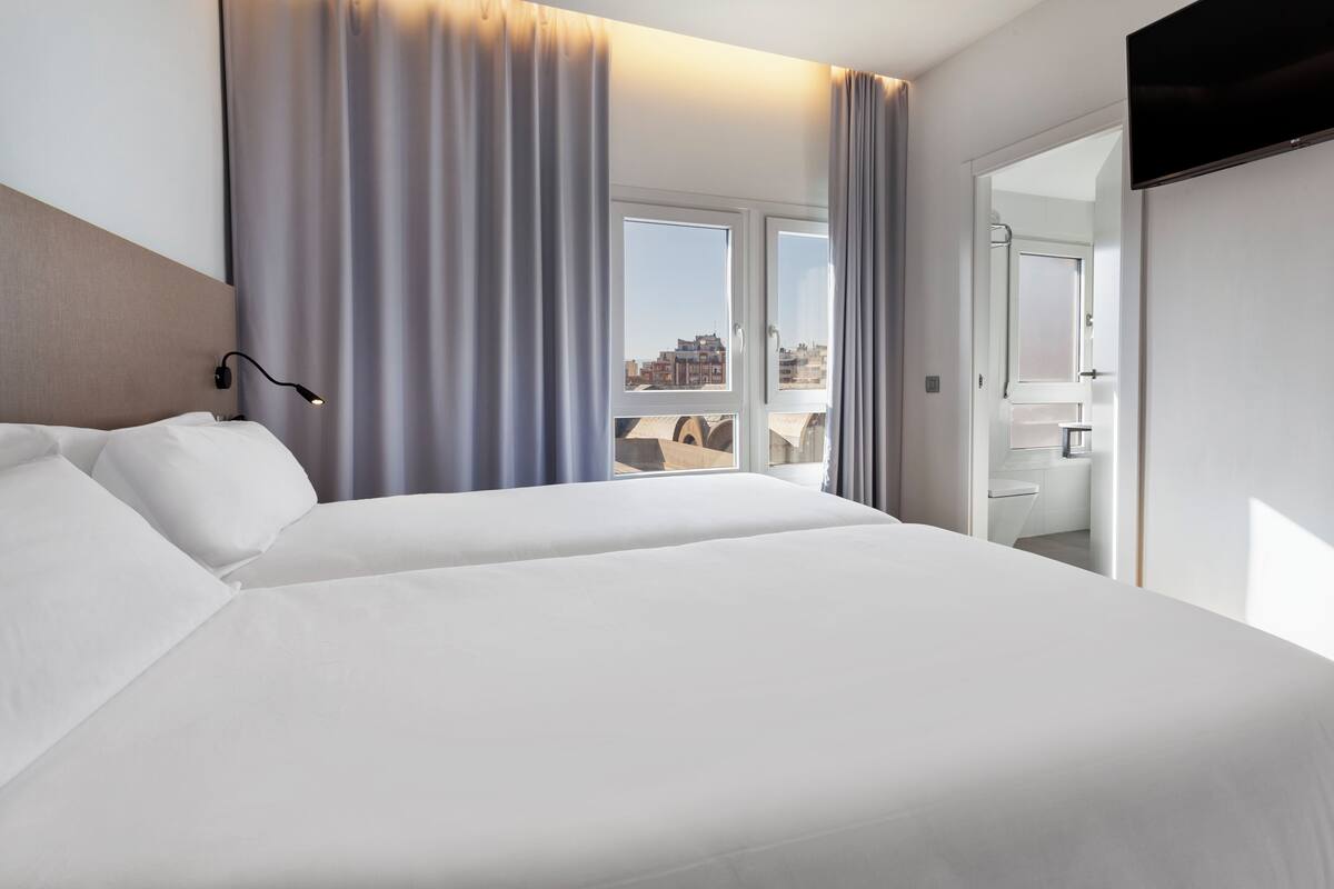 Une chambre d’hôtel moderne dotée d’un grand lit, d’un téléviseur à écran plat et offrant une vue sur la ville grâce à de grandes fenêtres.