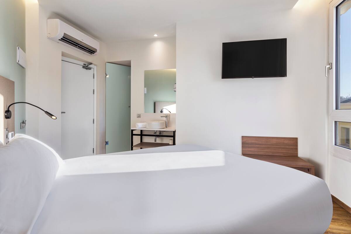 Une chambre d’hôtel moderne dotée d’un grand lit, d’un téléviseur à écran plat fixé au mur et d’une salle de bain visible à travers une porte ouverte.