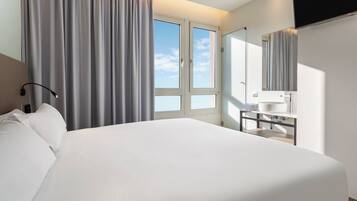 A modern hotel room with a large bed, a bedside table with a lamp, a bathroom with a mirror, and a window with curtains.