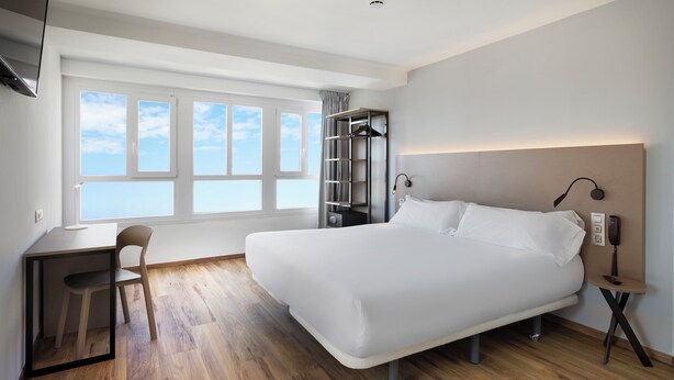 Habitació d'hotel moderna amb un llit gran, una taula de treball i una cadira. Hi ha una finestra amb vista al cel i un armari penjant.