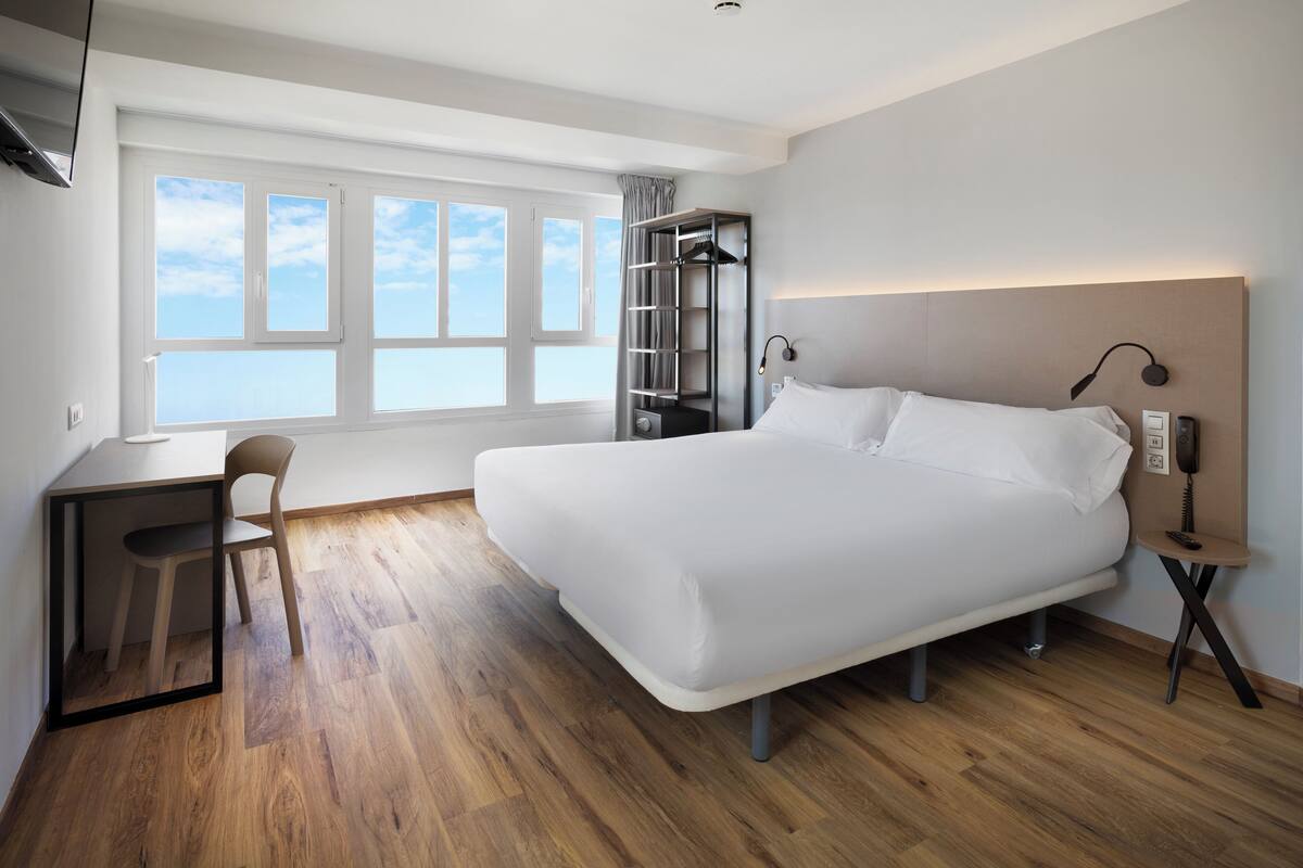 Une chambre d’hôtel moderne avec un grand lit, un bureau et une chaise. Il y a une fenêtre donnant sur le ciel et un placard suspendu.