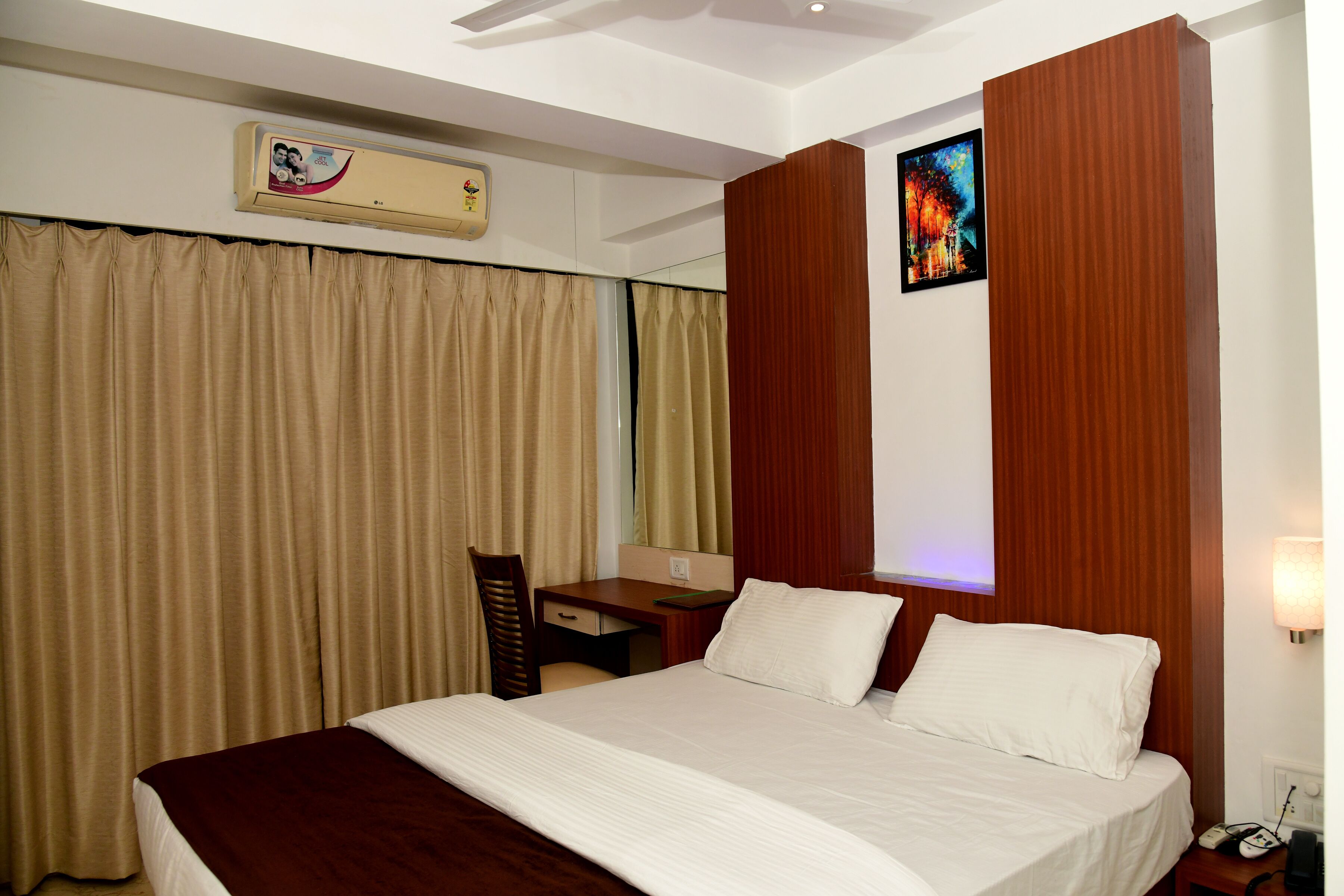 A hotel room with a bed, a desk, a chair, an air conditioner, and a painting on the wall.