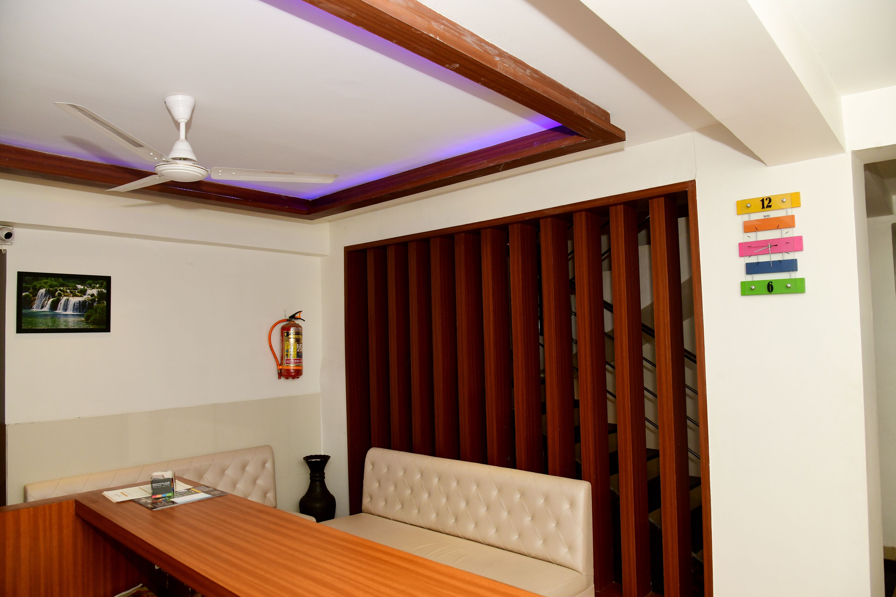 Una habitación con ventilador de techo, un banco de madera, una mesita con una revista, un reloj en la pared y un extintor.