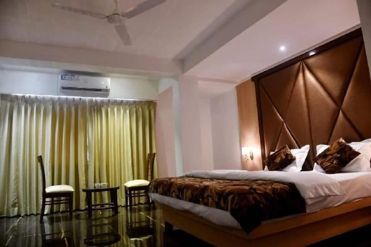 A hotel room with a bed, a ceiling fan, an air conditioning unit, a small table with chairs, and a window with curtains.