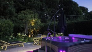Un bain à remous avec un éclairage violet, installé sur une terrasse en bois, entouré de verdure et agrémenté d’une petite table avec des chaises.