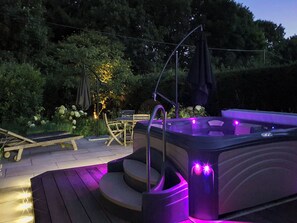 A hot tub with purple lighting on a wooden deck, surrounded by greenery and a small table with chairs.