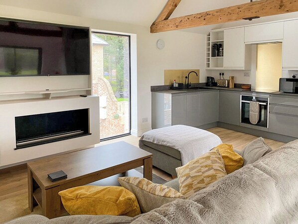 Un salon moderne avec un téléviseur, un canapé et une table basse. On aperçoit une cuisine avec des meubles gris et une fenêtre.