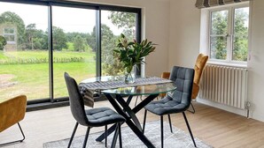 Un coin repas avec une table en verre, deux chaises, un tapis et une vue sur un paysage verdoyant grâce à de grandes fenêtres.