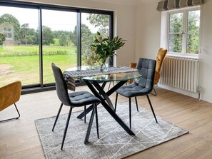 A dining area with a glass table, two chairs, a rug, and a view of a green landscape through large windows.