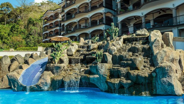 A hotel with multiple balconies, a pool with a waterfall feature, and a rocky landscape. - New listing! Stunning views. Fifth floor apartment (Punta Leona)