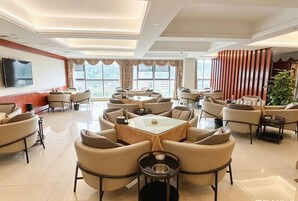 A hotel lobby with multiple seating areas, a flat-screen TV mounted on the wall, and large windows providing natural light.