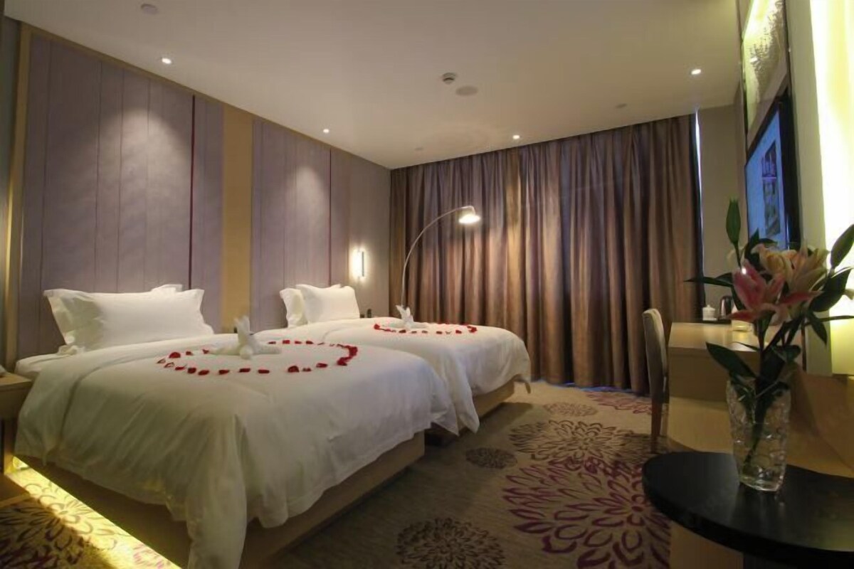 A hotel room with two beds, a large window with curtains, a vase with flowers, and a carpet with a floral pattern.