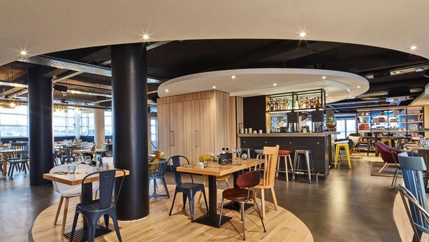 A modern restaurant with round tables, black and wooden chairs, a bar area, and a view of the parking lot. - Radisson Hotel Paris Le Bourget (Bonneuil-en-France)