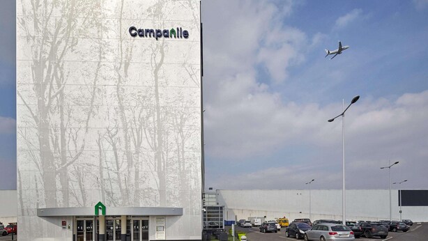A large building with a tree design on its facade, a parking lot with cars, and an airplane in the sky. - Radisson Hotel Paris Le Bourget (Bonneuil-en-France)