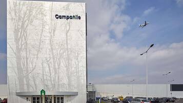 Un grand bâtiment dont la façade est ornée d’un motif d’arbre, un parking rempli de voitures et un avion dans le ciel.