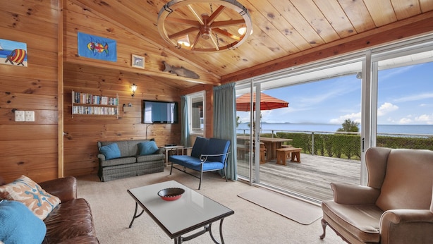 A cozy cabin interior with wooden walls, a television, a bookshelf, a dining area with a table and chairs, and a large sliding glass door leading to a deck with a view of the ocean.