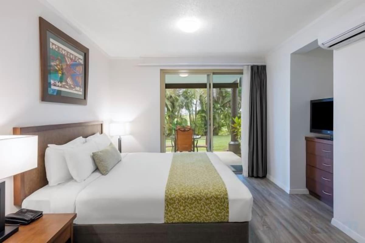 Habitació d'hotel amb llit, televisió, escriptori i balcó amb vista a vegetació.