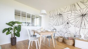 Un coin repas avec une table en bois, des chaises blanches, une plante en pot et un mur orné d’un motif abstrait en noir et blanc.