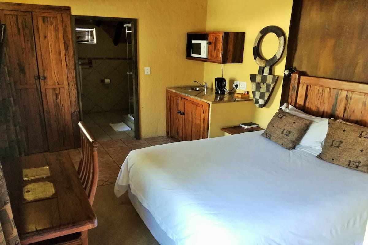 Een slaapkamer met een groot bed, een houten garderobe, een kleine kitchenette en een zichtbare badkamer door een open deur.
