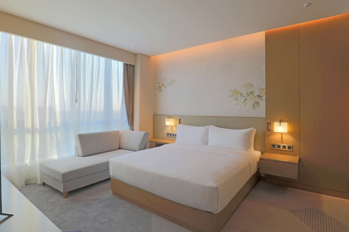 A hotel room with a large bed, a sofa, two bedside lamps, a nightstand, and a large window with sheer curtains.