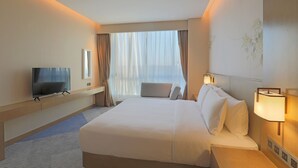 Habitación de hotel con una cama grande, un televisor de pantalla plana, una mesita de noche y una ventana con cortinas translúcidas.