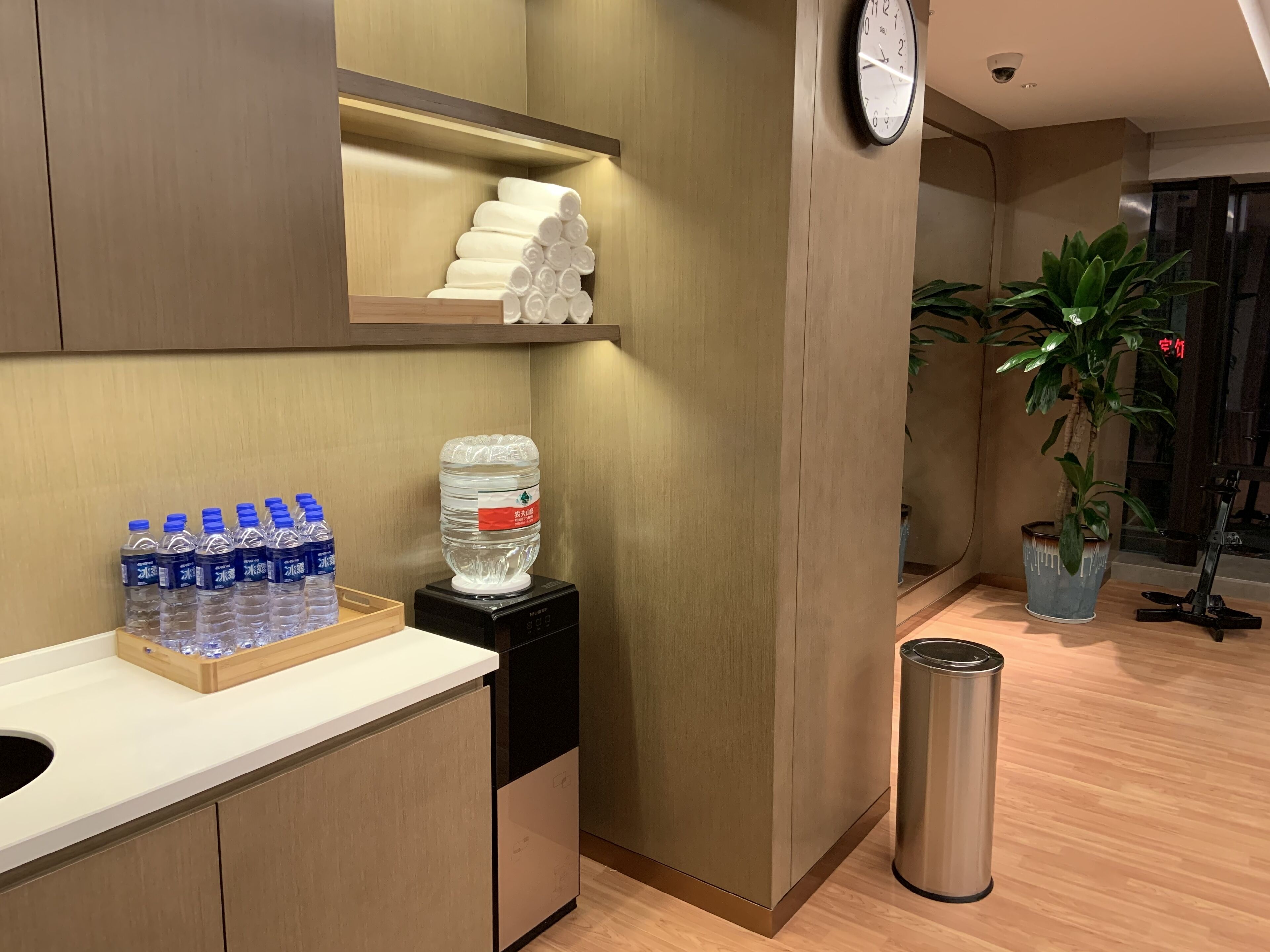 A hotel reception area with a water dispenser, bottled water, a large water jug, a clock, and a potted plant.