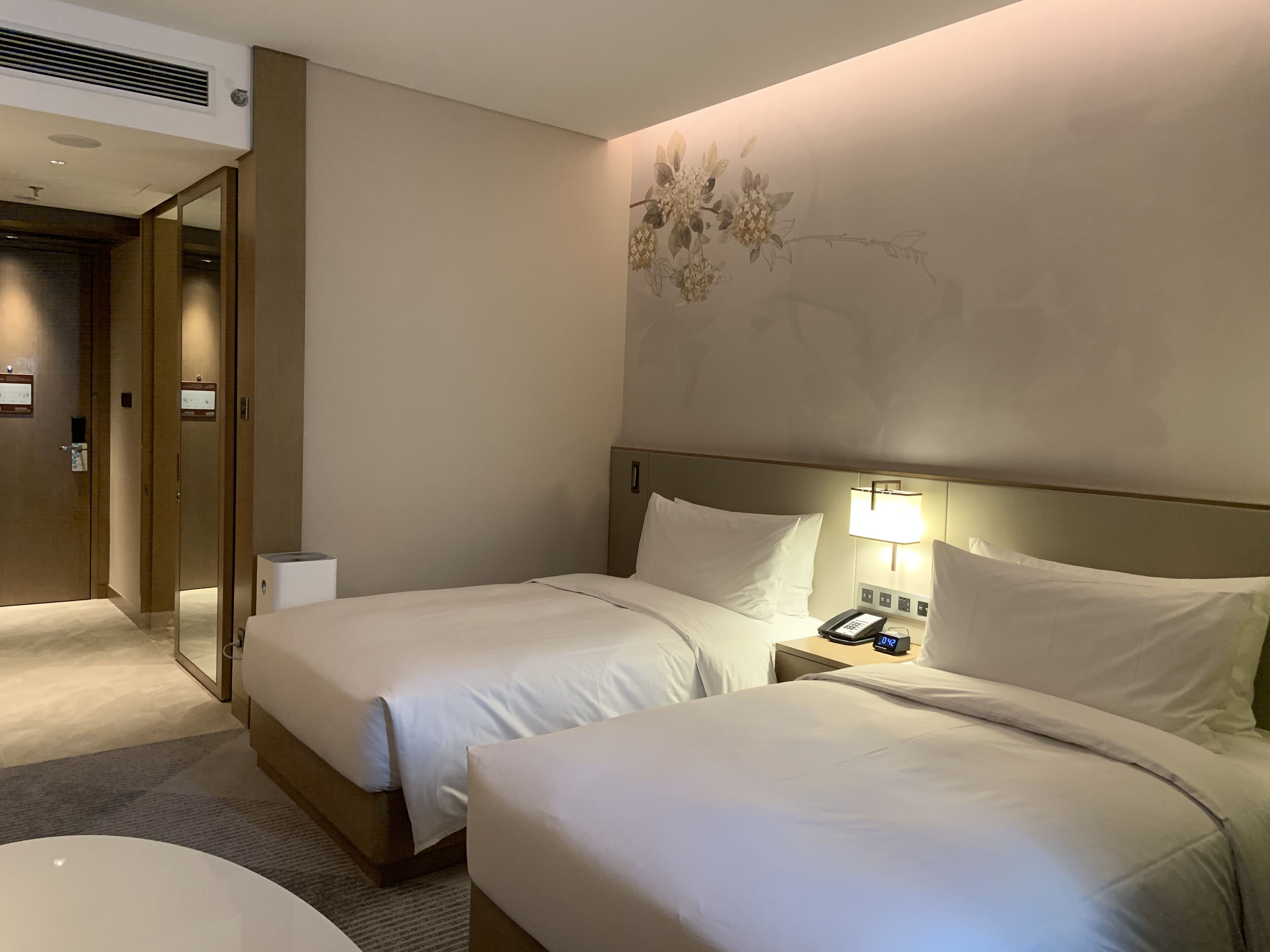 A hotel room with two beds, a bedside table with a lamp, a phone, and a wall-mounted air conditioning unit.