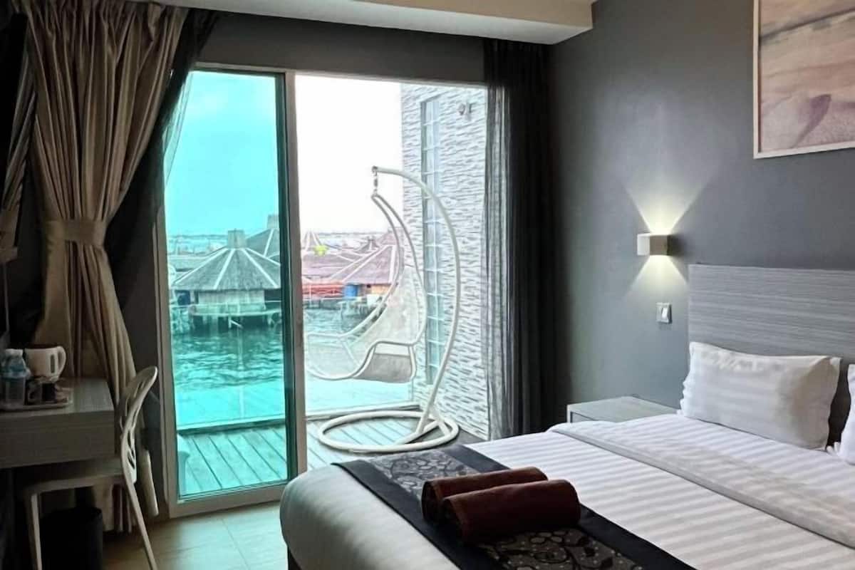A hotel room with a bed, a desk with a chair, a large window with a view of a water body and a bridge, and a wall-mounted light fixture.