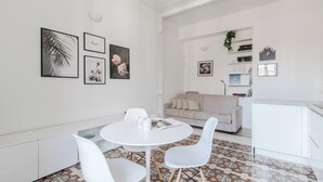 Un salon moderne et bien éclairé, avec une table à manger ronde, des chaises blanches, un canapé beige et un coin cuisine doté de meubles blancs et d’une horloge au mur.