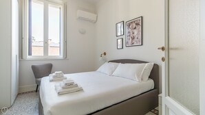 Un lit bien fait, avec du linge de lit et des serviettes blanches, une chaise grise, une fenêtre avec des stores et deux tableaux encadrés accrochés au mur.