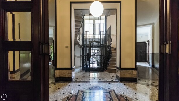 Un couloir avec un sol carrelé, un escalier et un portail en métal noir.