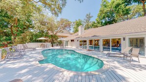 Ein Schwimmbecken mit Holzterrasse, Liegestühlen und einem überdachten Bereich mit Glasüren.