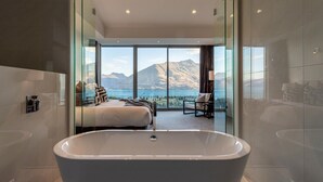Een moderne badkamer met een vrijstaand bad, een bed en een zithoek met uitzicht op bergen en een meer.