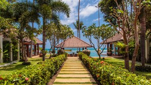 Uma passagem leva a uma área de piscina rodeada por vegetação exuberante e palmeiras, com uma vista clara do oceano ao fundo.