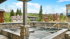 A stone hot tub with a metal ladder, surrounded by a stone wall, set against a backdrop of buildings and trees.