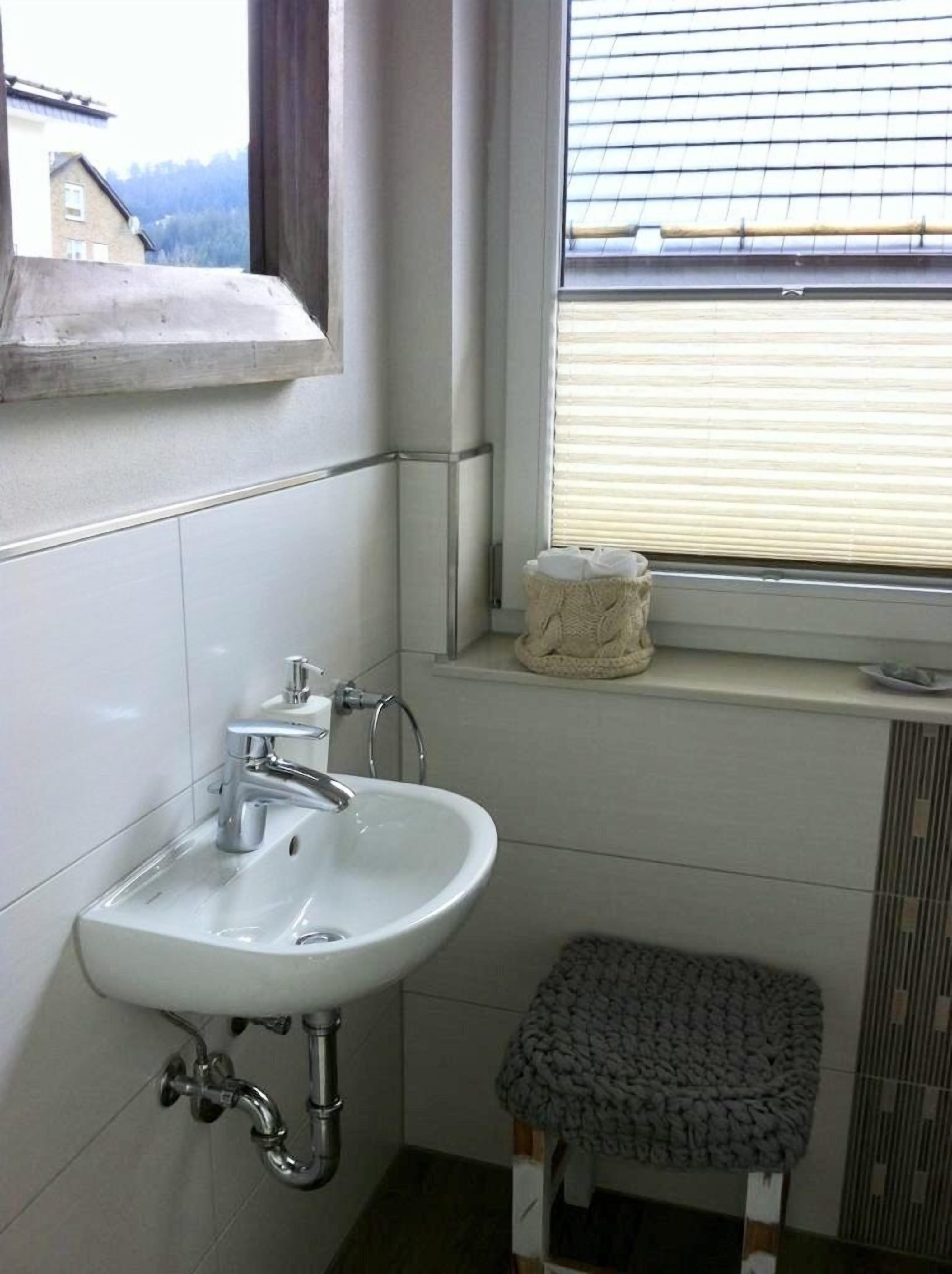 A bathroom with a white sink, a window with blinds, and a small stool.