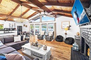 A spacious living area with a high ceiling, wooden beams, a fireplace, and a large window overlooking a lake.