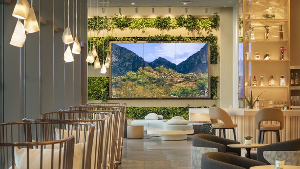 A modern interior with a large wall-mounted TV displaying a mountainous landscape, seating areas with cushioned chairs and tables, and a bar area with shelves of bottles and glasses. - The Westin Tempe (Tempe)