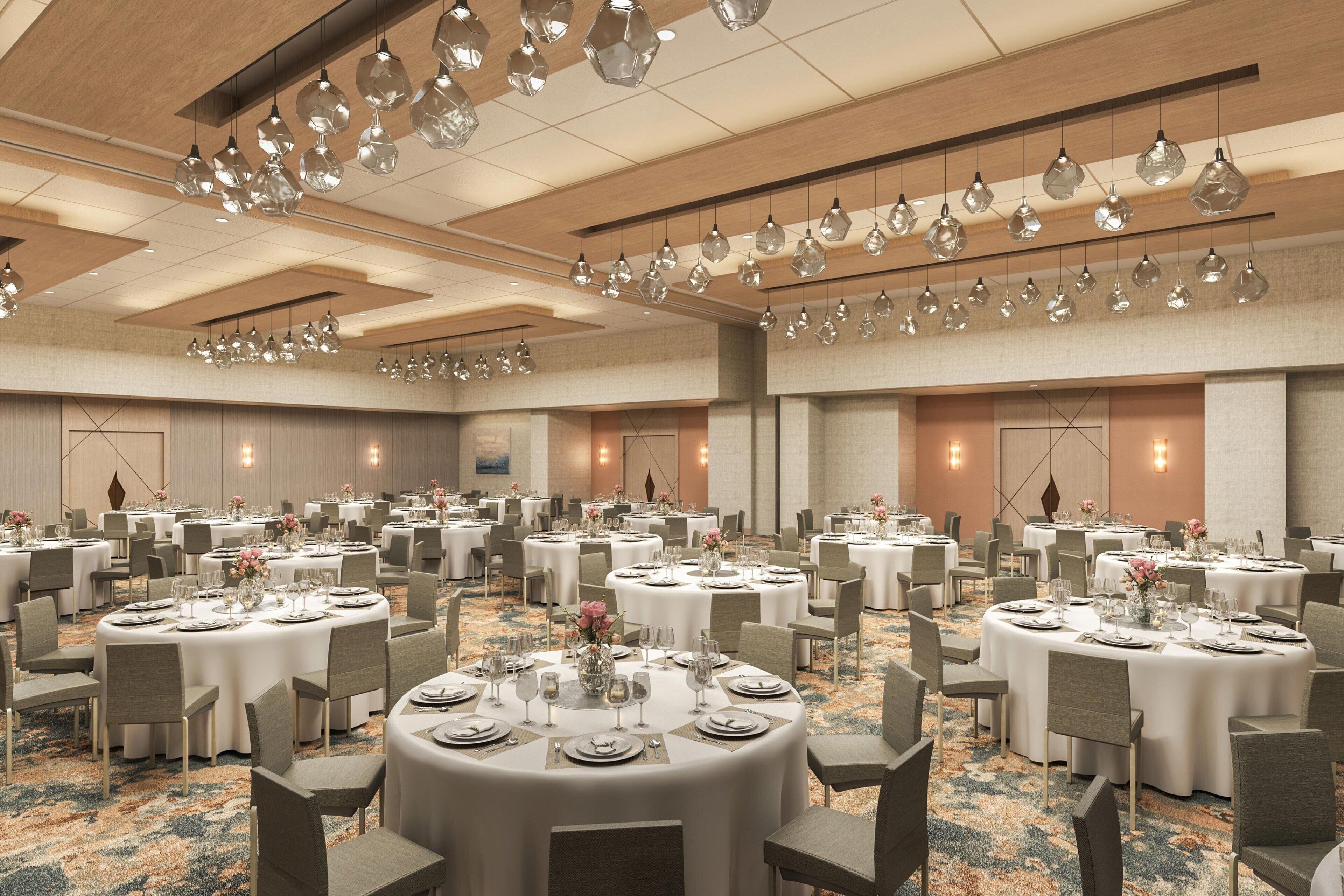 A large banquet hall with round tables set for dining, featuring a modern chandelier and neutral color scheme.