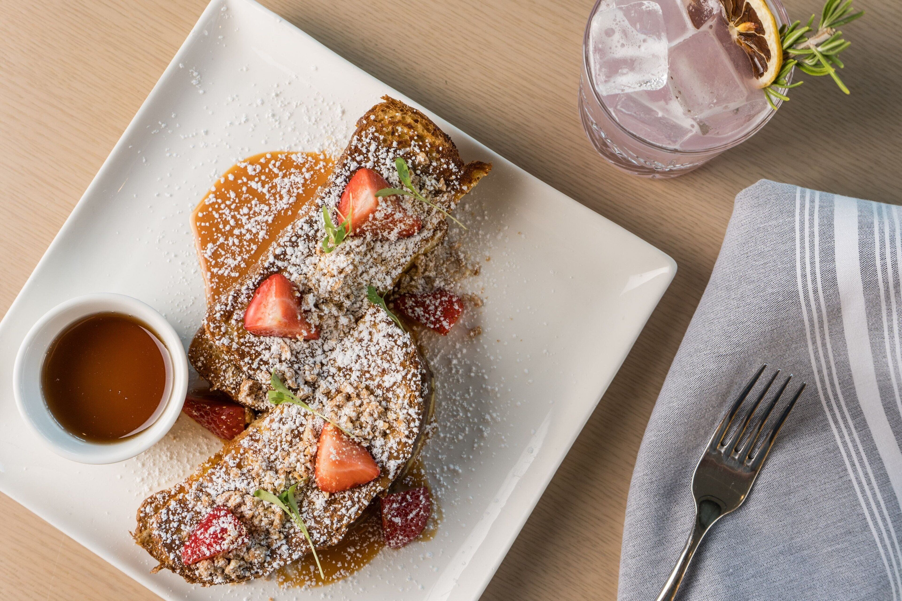 Une assiette de pain perdu garnie de fraises et saupoudrée de sucre glace, un verre de thé glacé avec une tranche de citron et une fourchette posée sur une serviette.