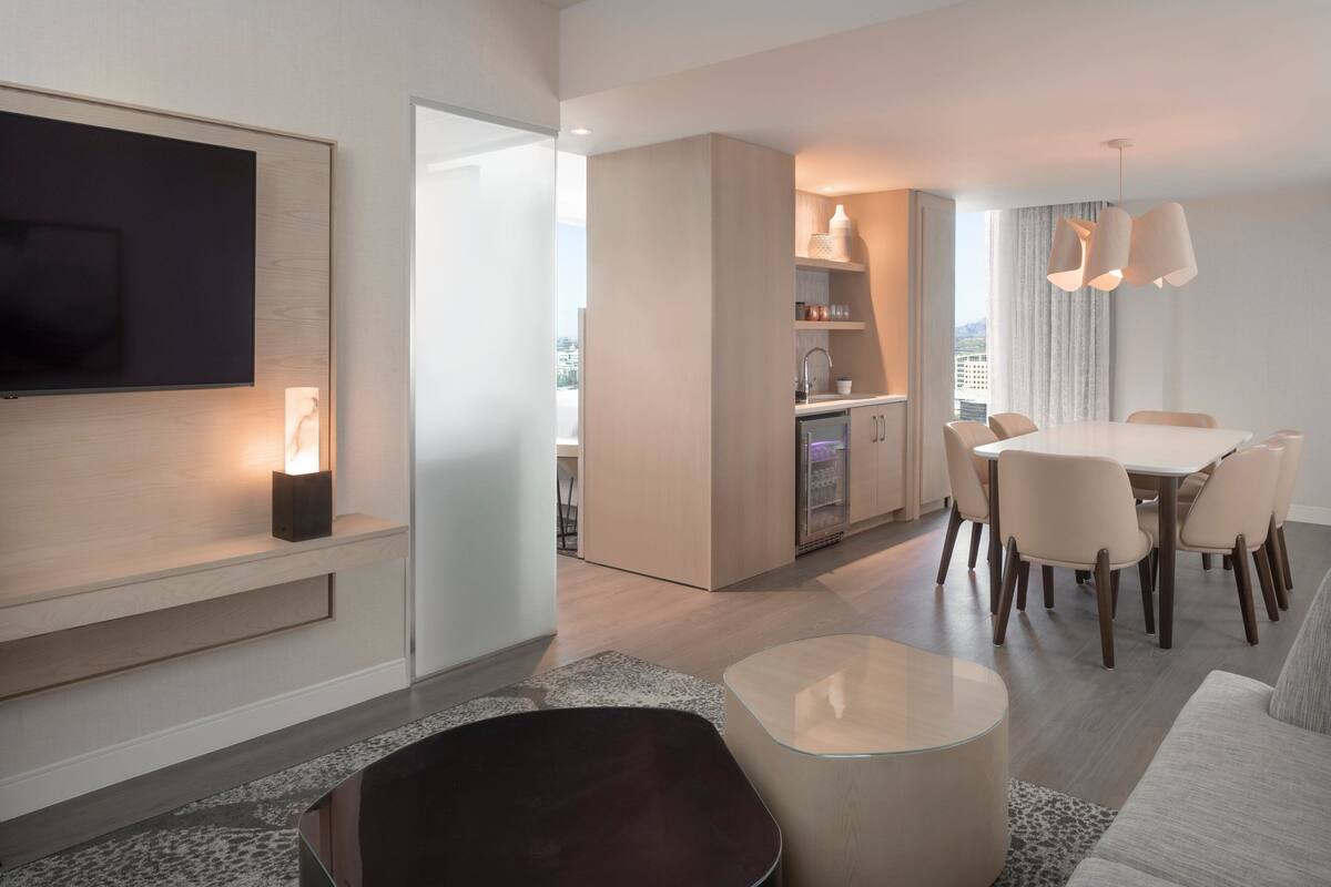 A modern hotel room with a flat-screen TV, a dining area with a table and chairs, a kitchen area, and a large window offering a city view.