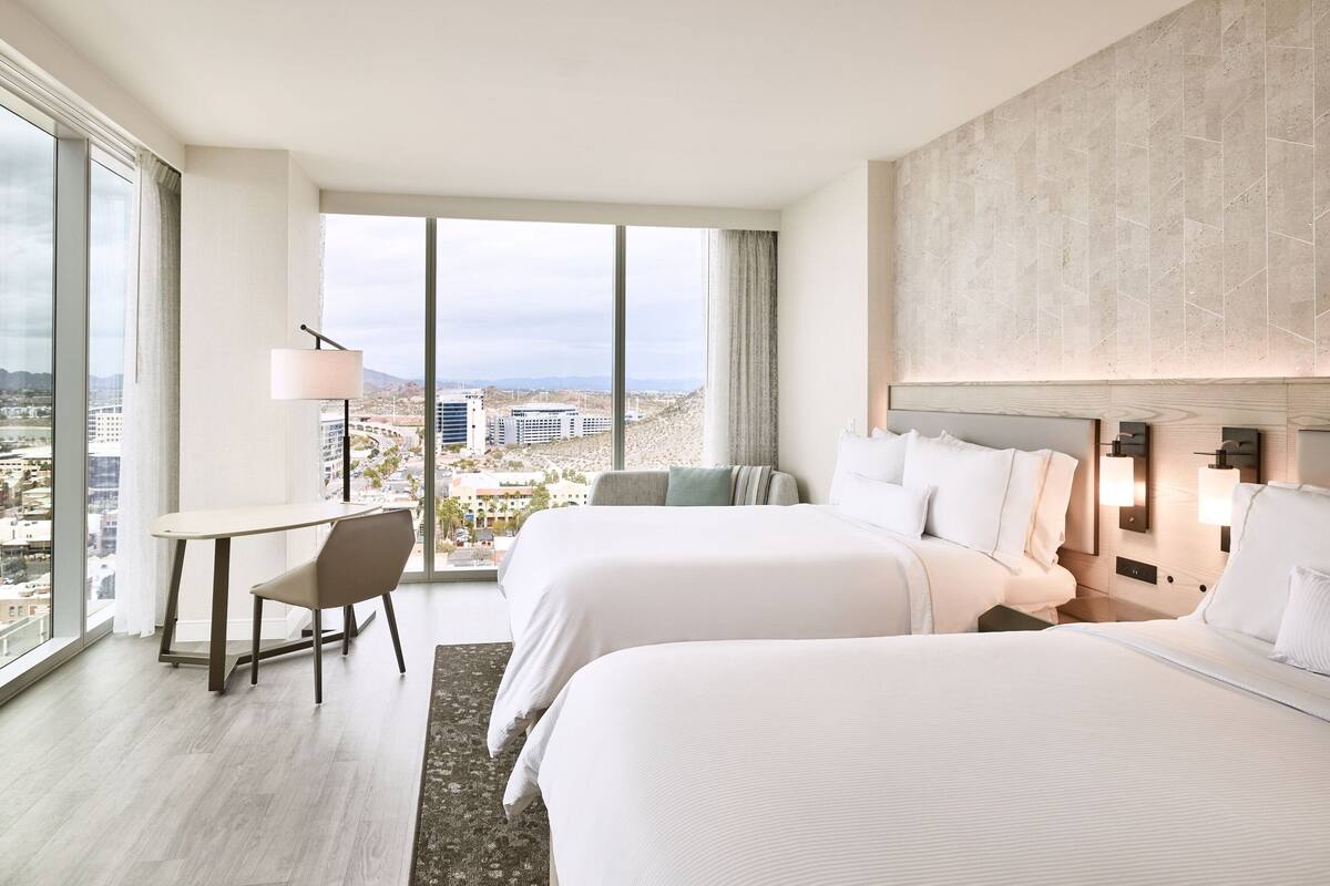 A hotel room with two beds, a desk, and a chair. There is a large window offering a city view.