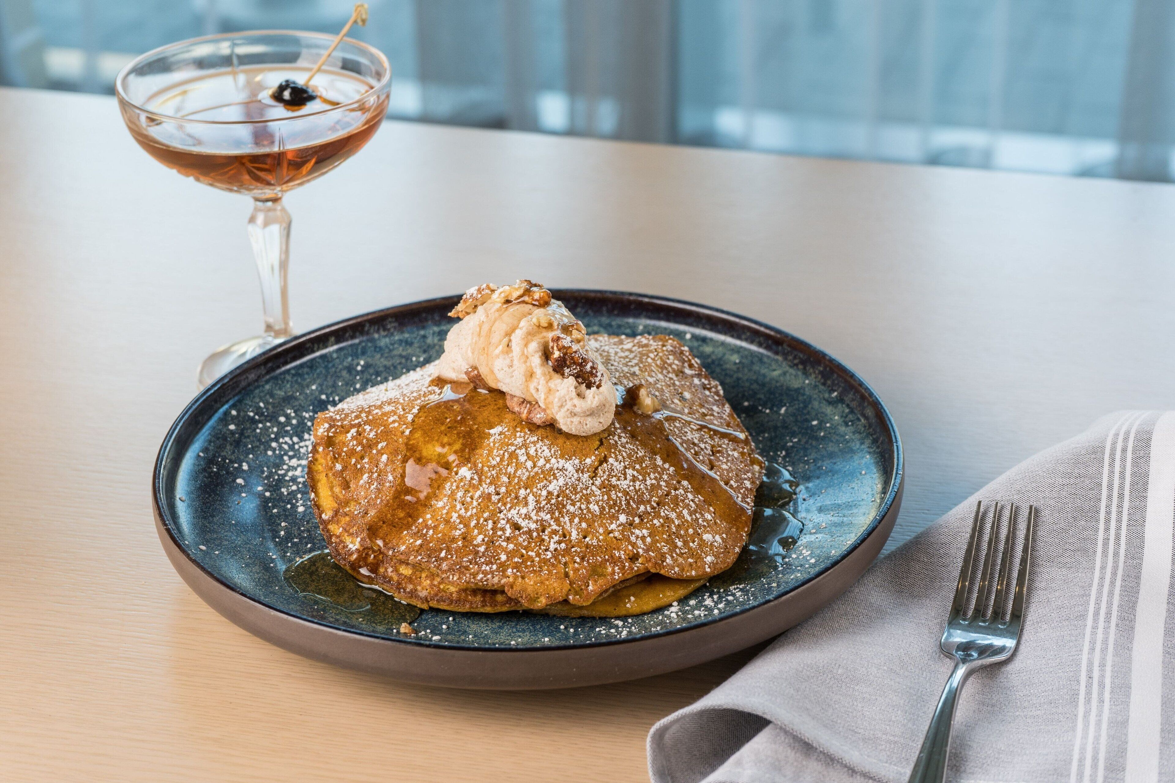 A dessert plate with a pancake topped with a dollop of ice cream and a sprinkle of powdered sugar, accompanied by a glass of a dark-colored beverage.