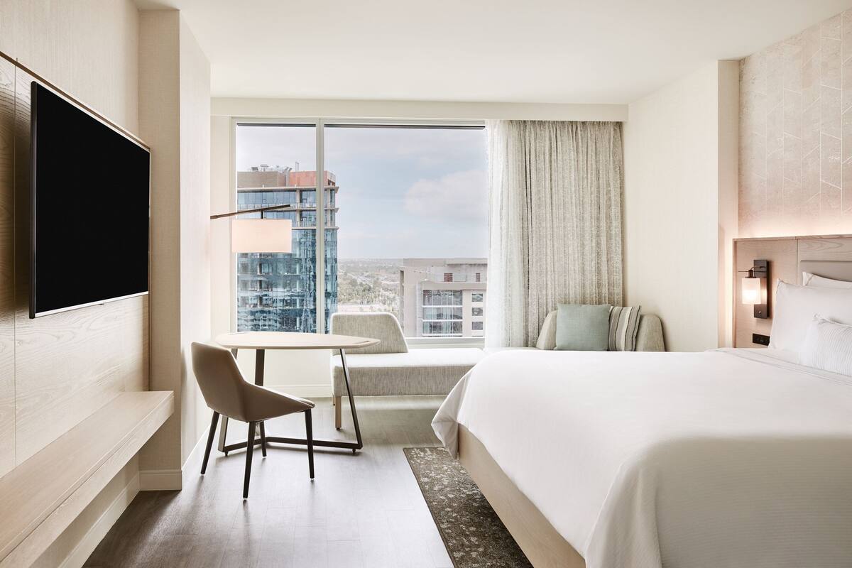A modern hotel room with a large bed, a flat-screen TV mounted on the wall, a small table, and a comfortable chair.