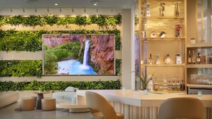 A modern bar area with a large TV screen displaying a waterfall, surrounded by greenery and bar shelves with bottles and glasses. - The Westin Tempe (Tempe)