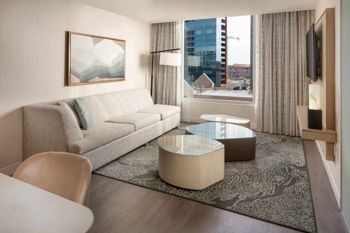 A modern hotel room with a sofa, two glass coffee tables, a large window with curtains, a flat-screen TV, and a framed abstract painting.