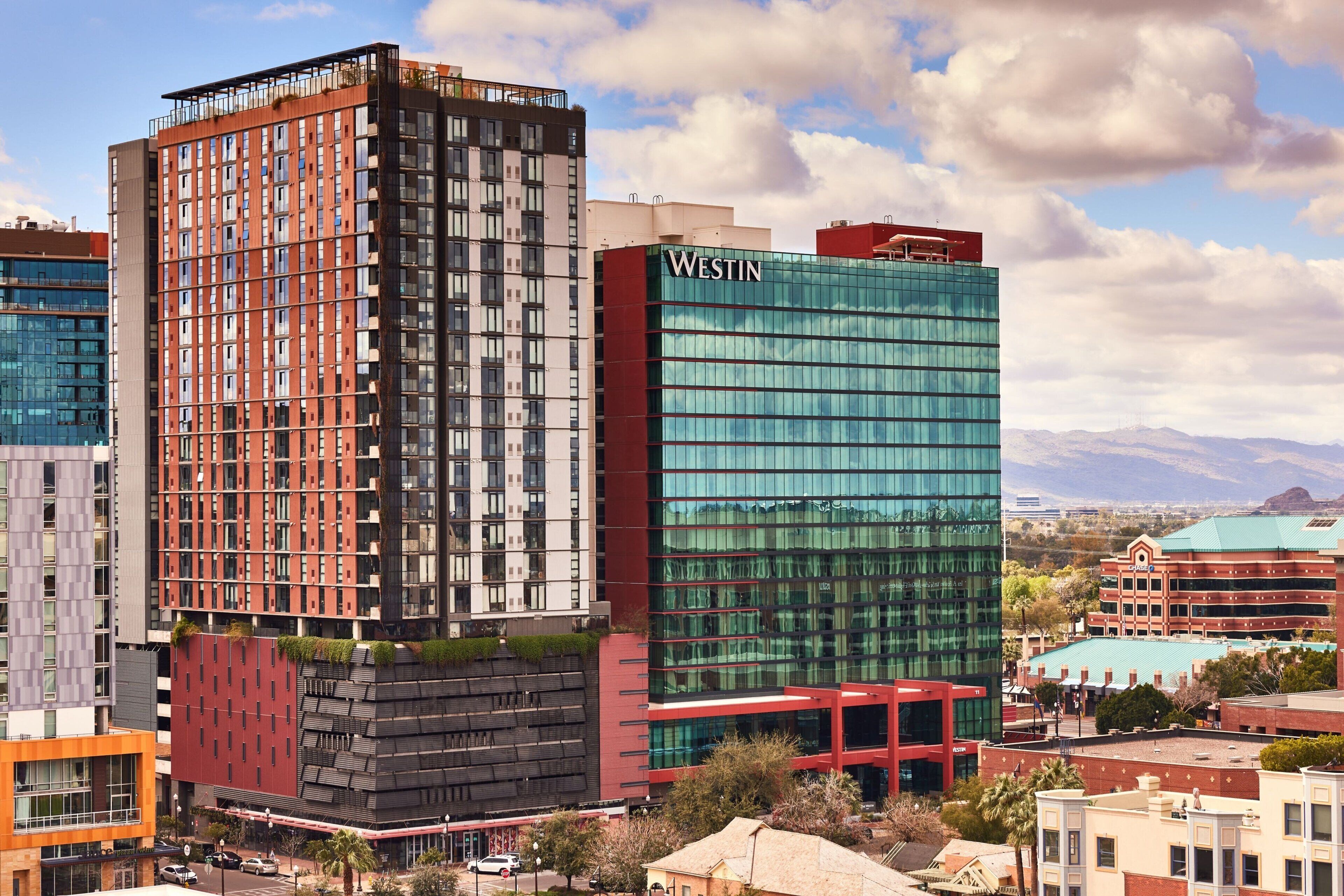 A modern hotel with a prominent 'Westin' sign, featuring a mix of glass and brick facades, and surrounded by other buildings and a mountainous backdrop.